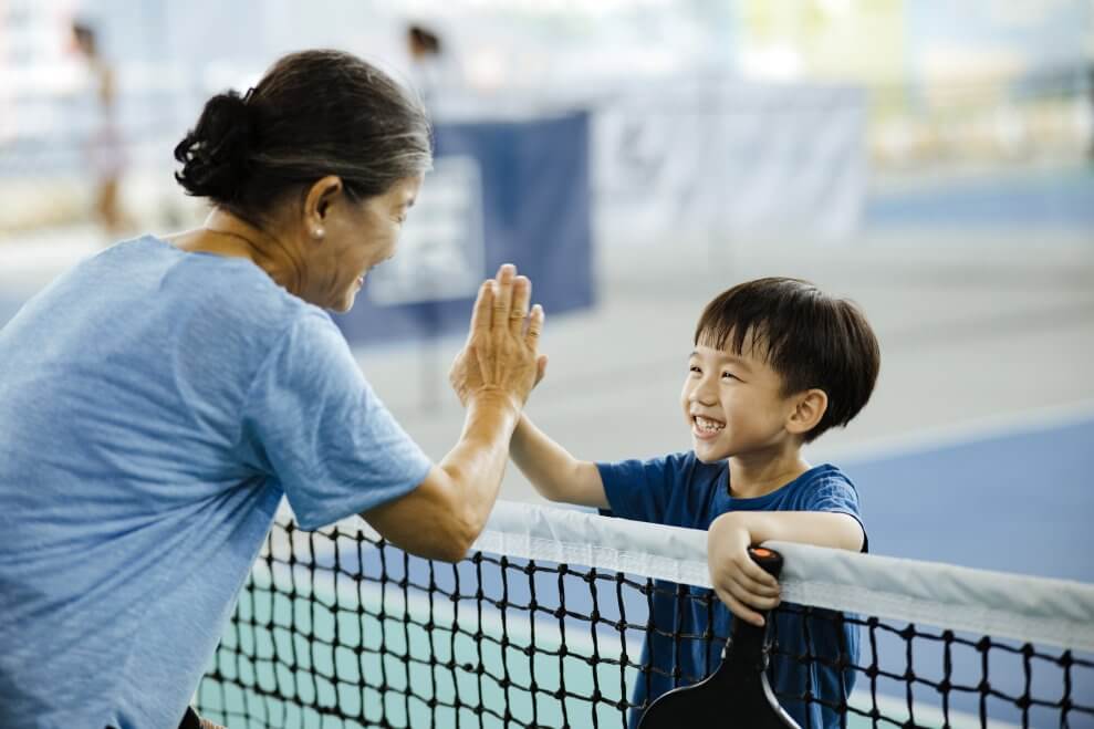 A young boy and his grandmother high five on the pickleball court as they smile at each other. The boy holds a pickleball paddle in his other hand.
