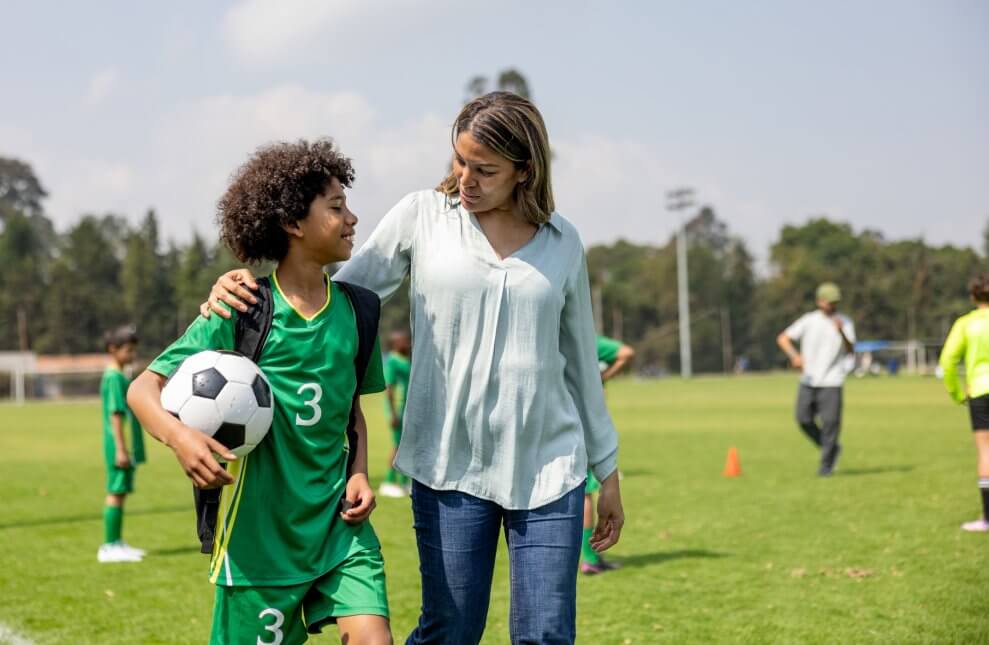 A mom walks across a soccer field with her preteen son, who's wearing a soccer uniform and carrying a soccer ball. She has her arm around his shoulder.