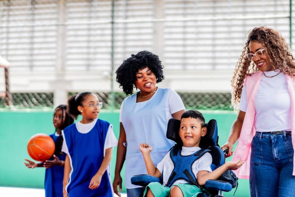 A group of students, including one student in a wheelchair, gather on a basketball court for phys ed class. Two teachers stand with the students.
