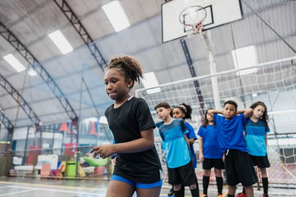 An elementary-age girl prepares to run drills in phys ed class. Her classmates wait in a line behind her.