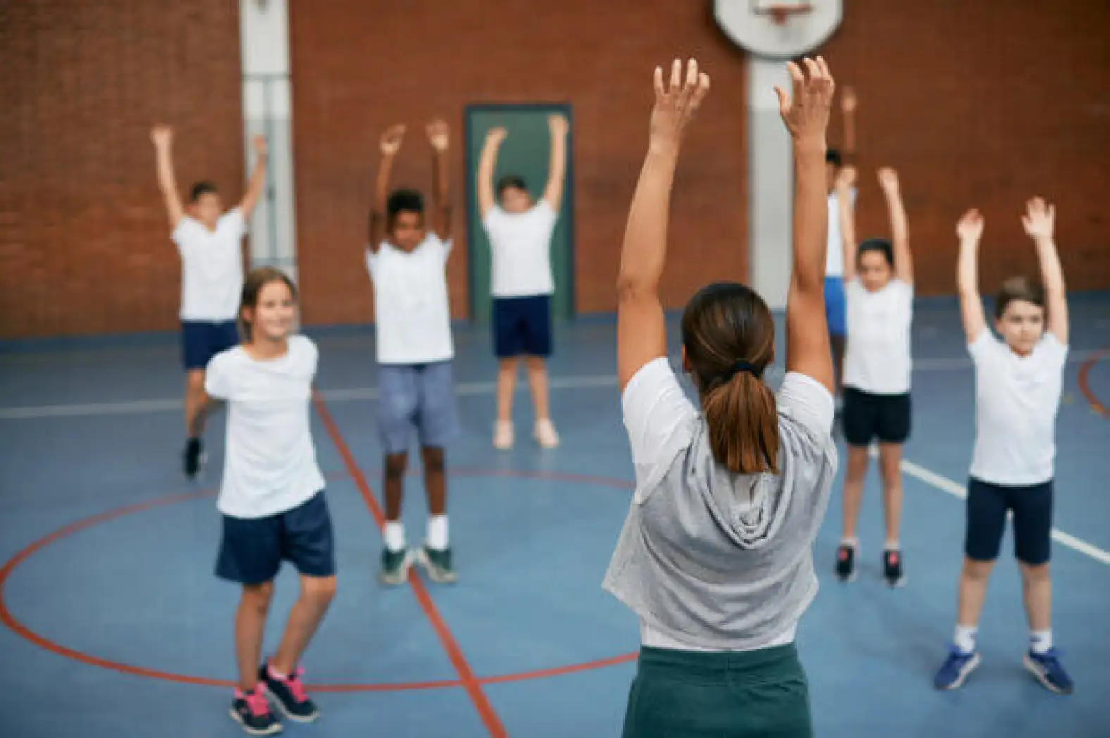 A gym teacher stands in front of her students and they all stretch with their arms over their heads