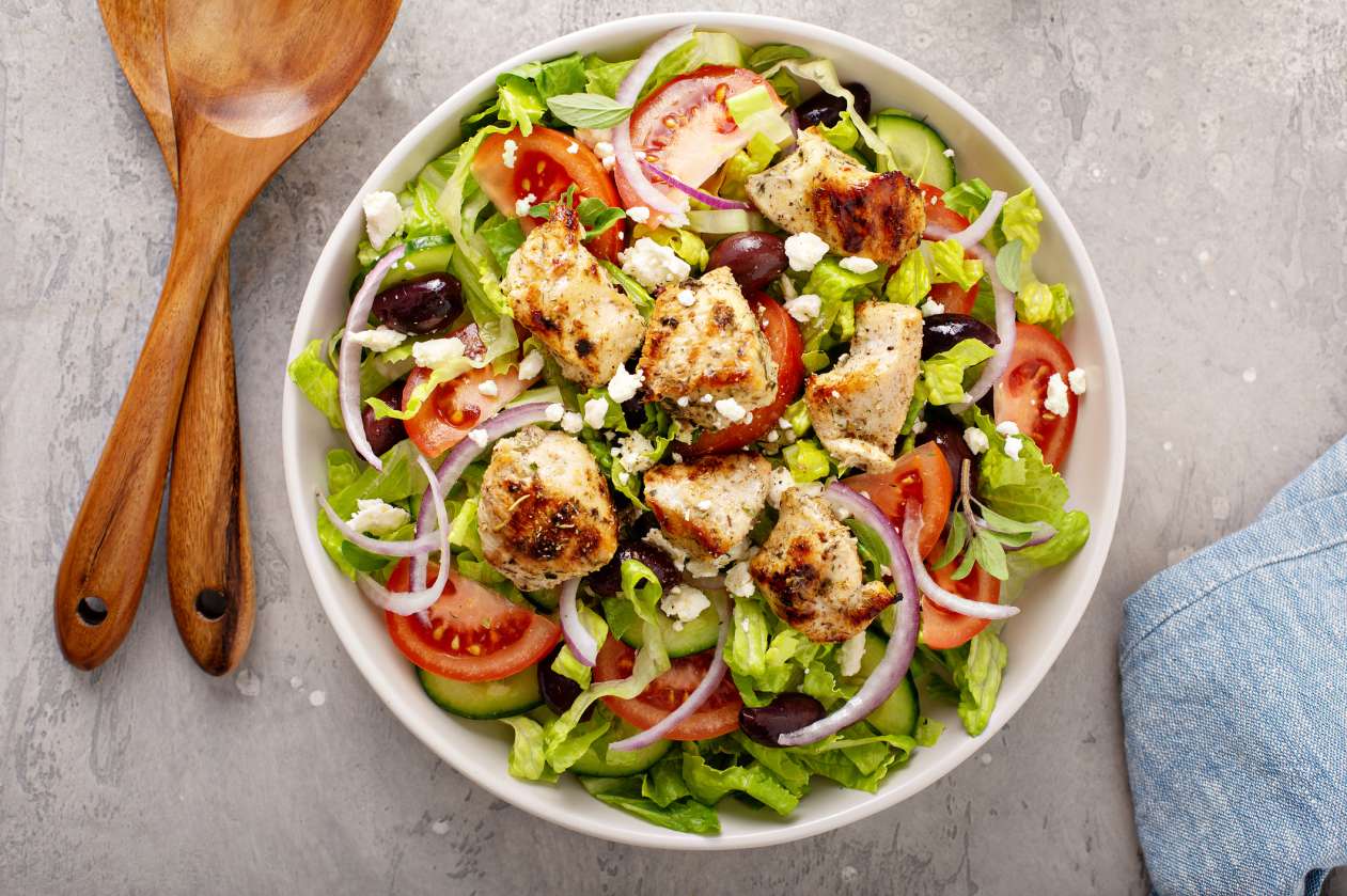 A bowl of colourful salad, featuring lettuce, olives, chicken, onions, and tomatoes.