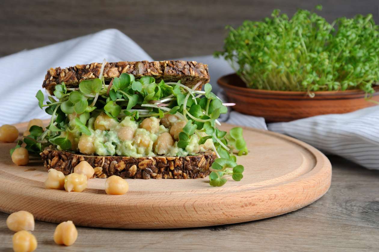 A sandwich packed full of greens, chickpeas, and avocado sits on a wooden platter.