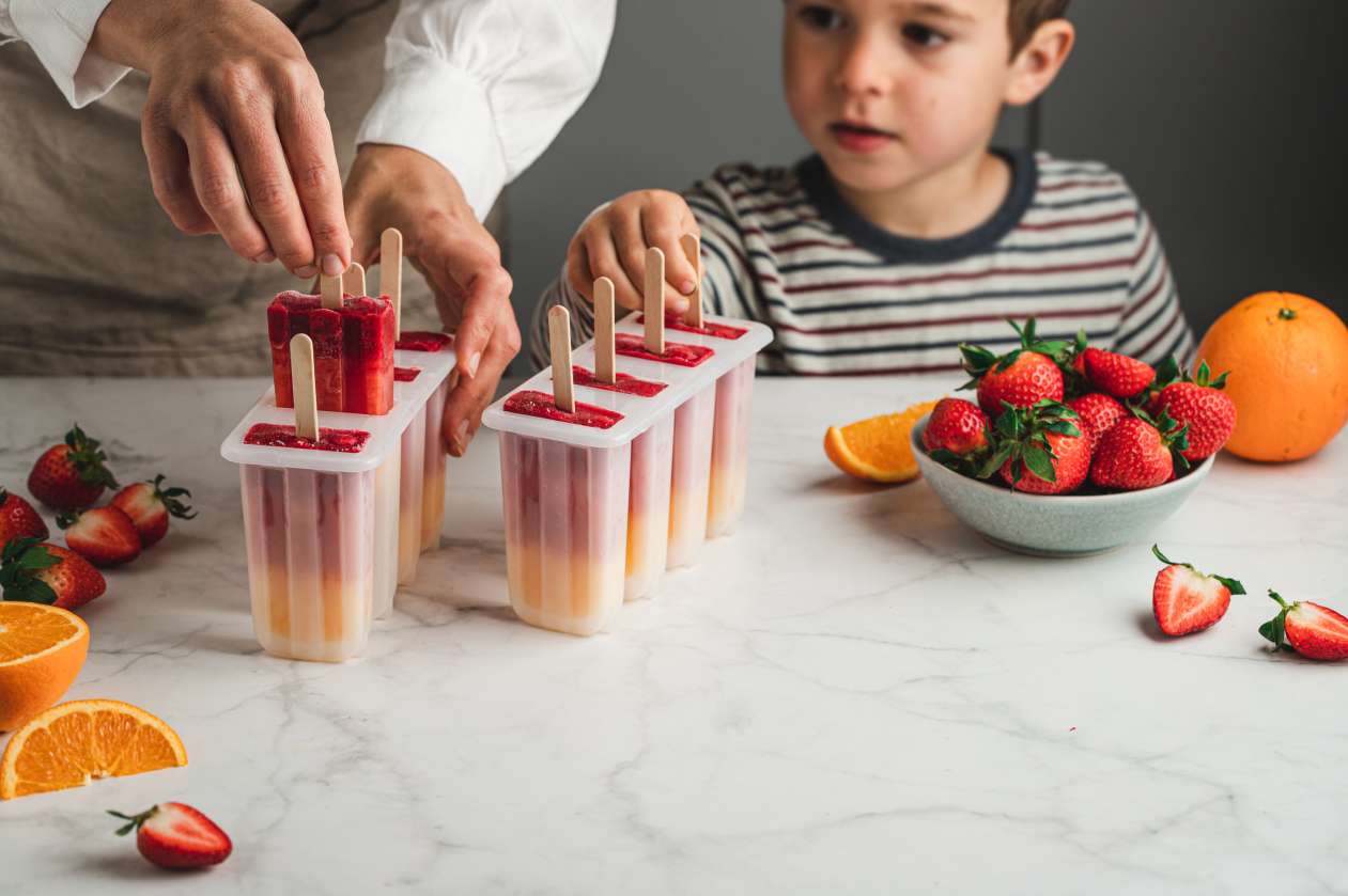 A child and his parent stand at the counter and help themselves to some homemade fruit popsicles.