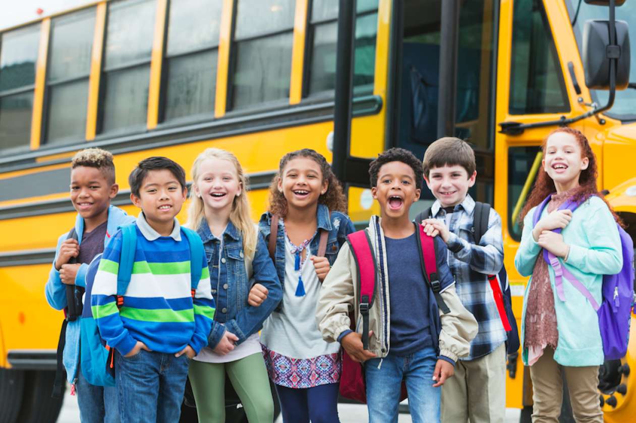 A group of smiling and laughing kids stand in front of a school bus.