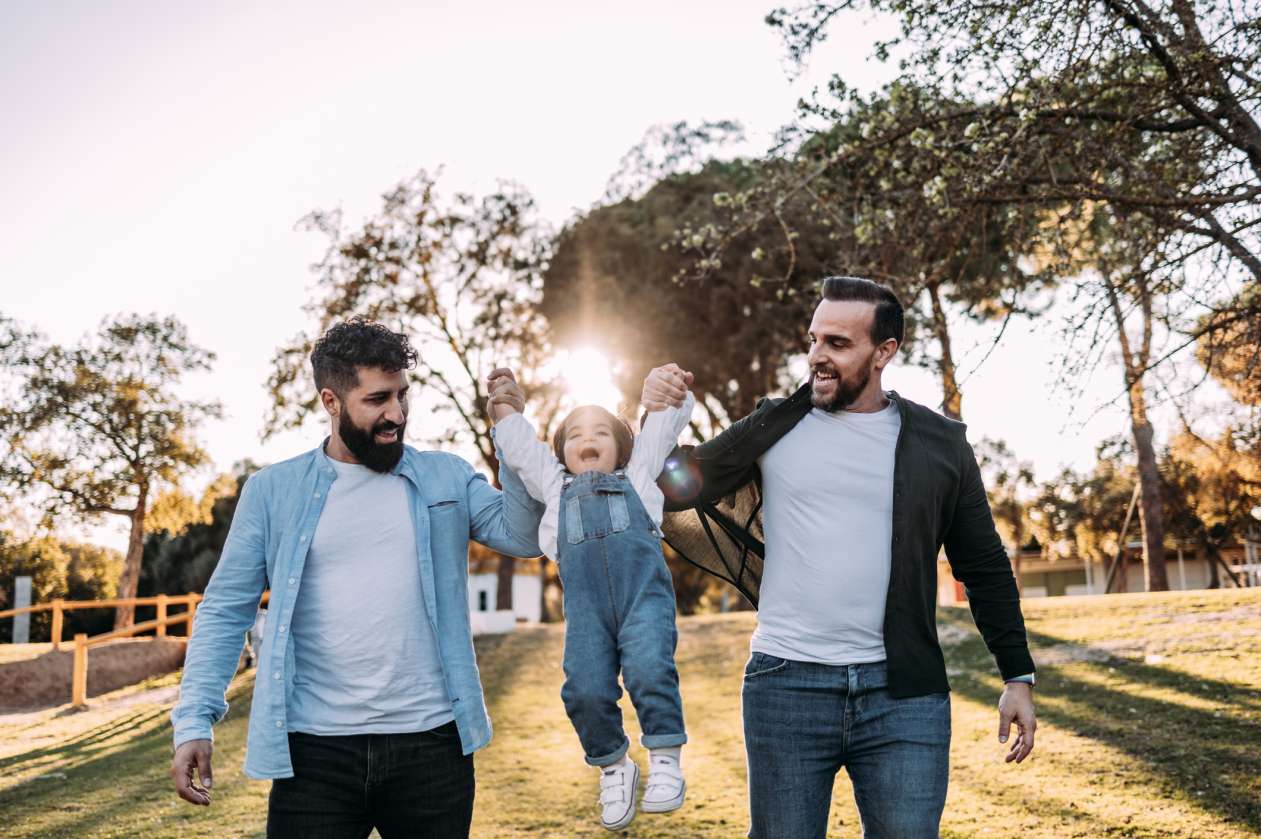 Two dads walk outside with their toddler daughter.