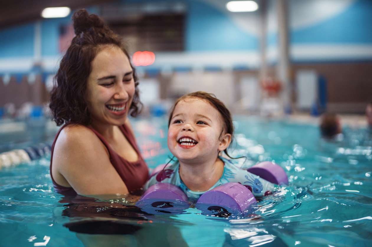 A mom supports her young toddler as she swims with floaties on her arms in an indoor swimming pool.