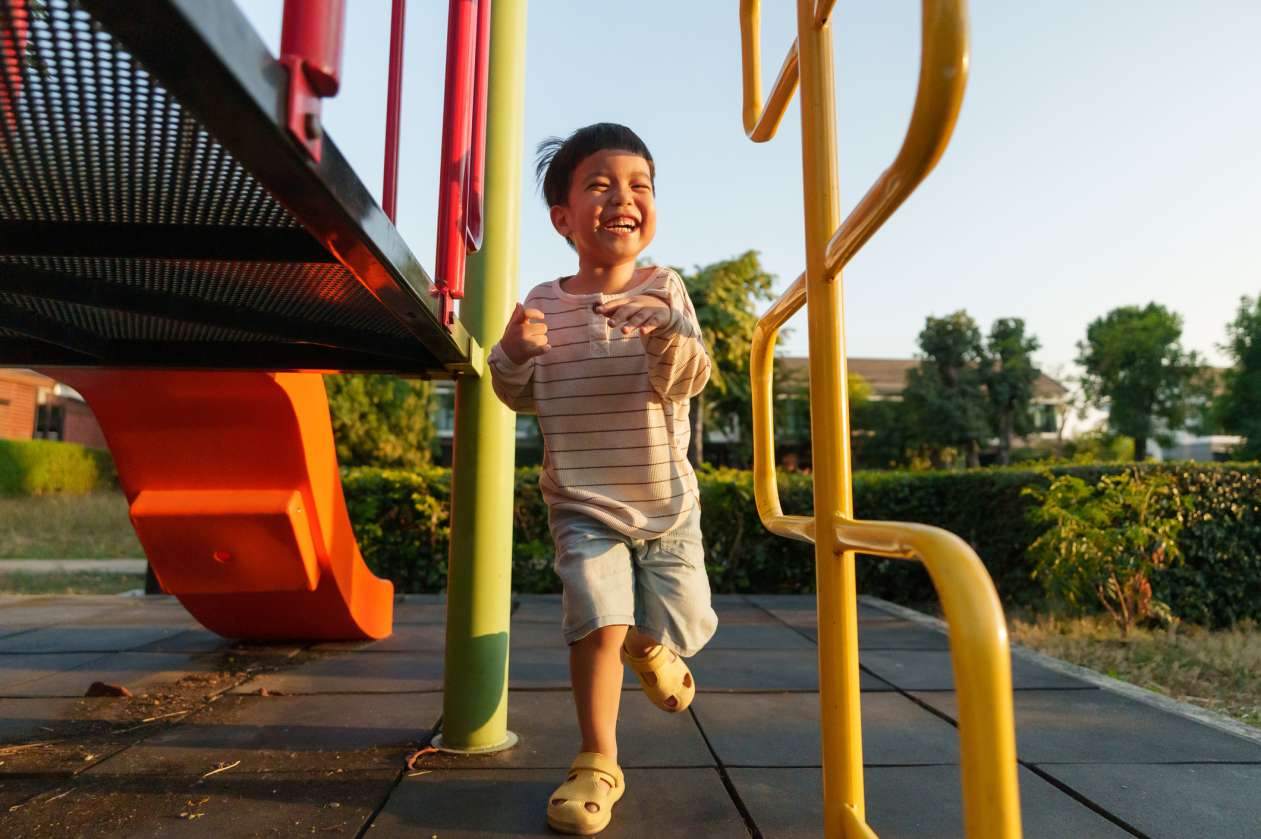 A young boy laughs as he runs around a playground on a sunny evening.
