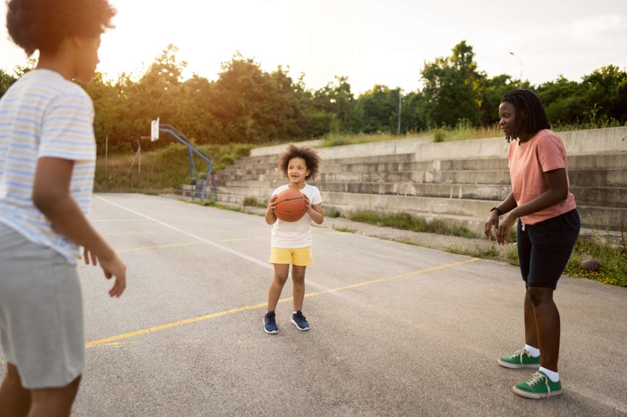 A mother and her two children play outside on a basketball court. The youngest child holds the ball and is about to pass it.