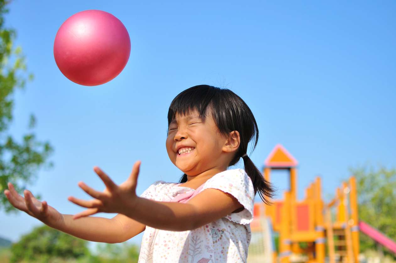 A young girl closes her eyes and smiles as she tries to catch a bouncy ball at the playground. 