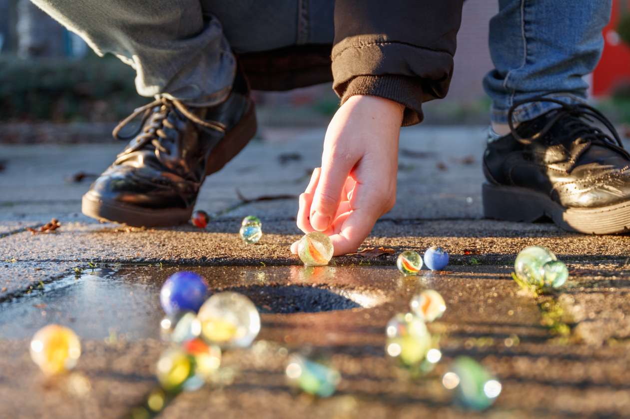 A child crouches down on the pavement outside on a sunny day and plays marbles