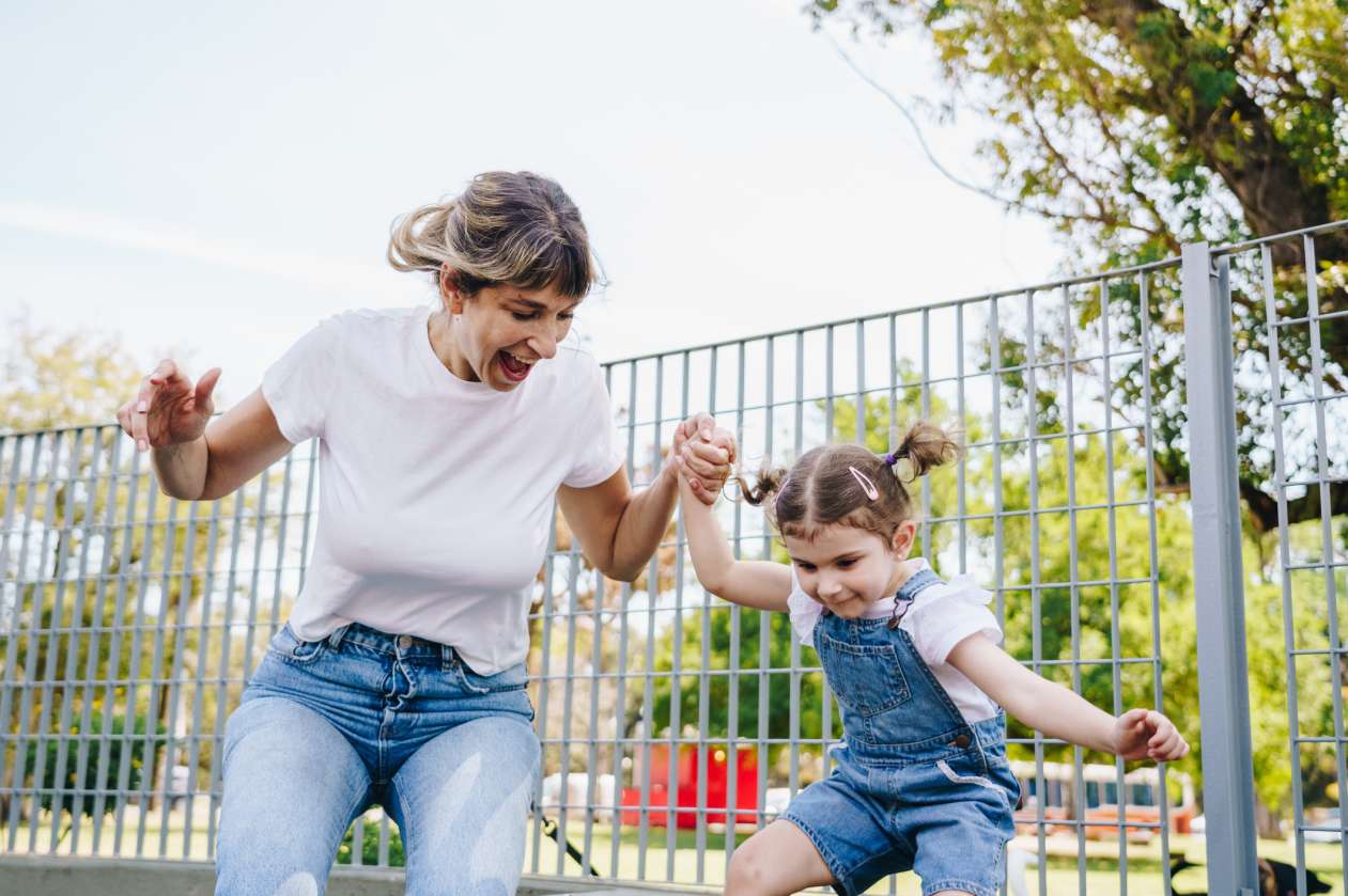 A mom and her daughter hold hands while they jump together outside at a park.