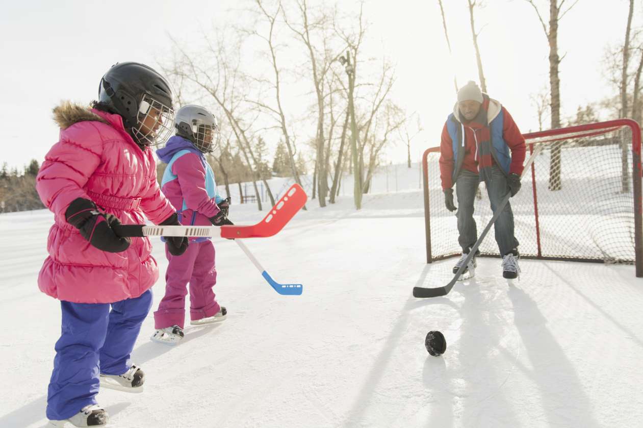 A father stands in front of a hockey net on an outdoor rink in the winter. His two young daughters stand on the ice in front of him, each holding a hockey stick.