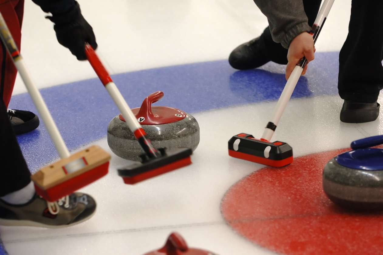 Curling players hold their brooms on the ice next to a curling stone.