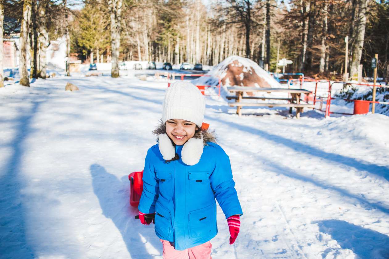 A girl pulls a sled through a park on a sunny winter day. She's smiling and squinting at the sun.