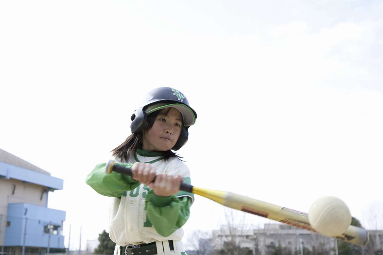 A girl wearing a baseball uniform and helmet hits a baseball with a bat.