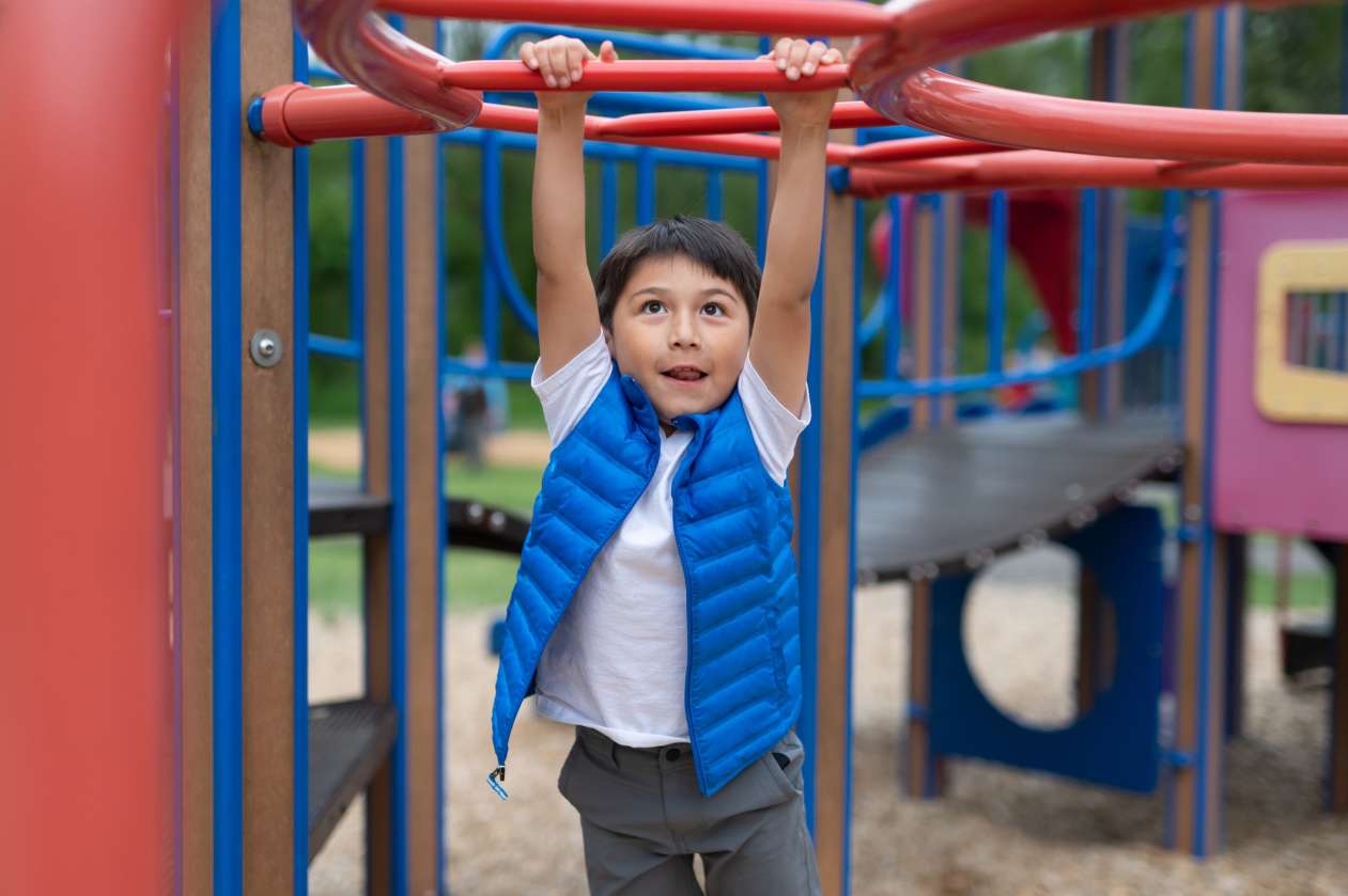 A young boy hangs from the monkey bars at a playground.