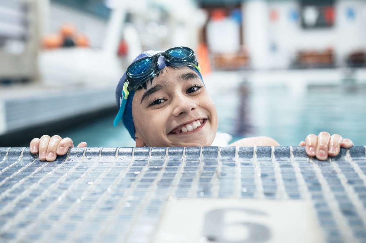 A boy wearing a swim cap and goggles grips the side of the pool, smiling. Swimming is the first event in a triathlon.