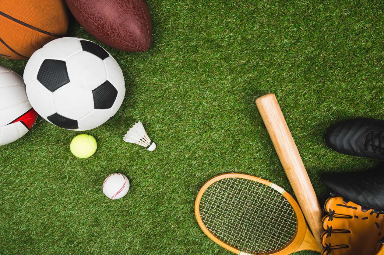 A pile of sports equipment, including balls, a baseball bat and glove, and a badminton racket, lies on the grass.