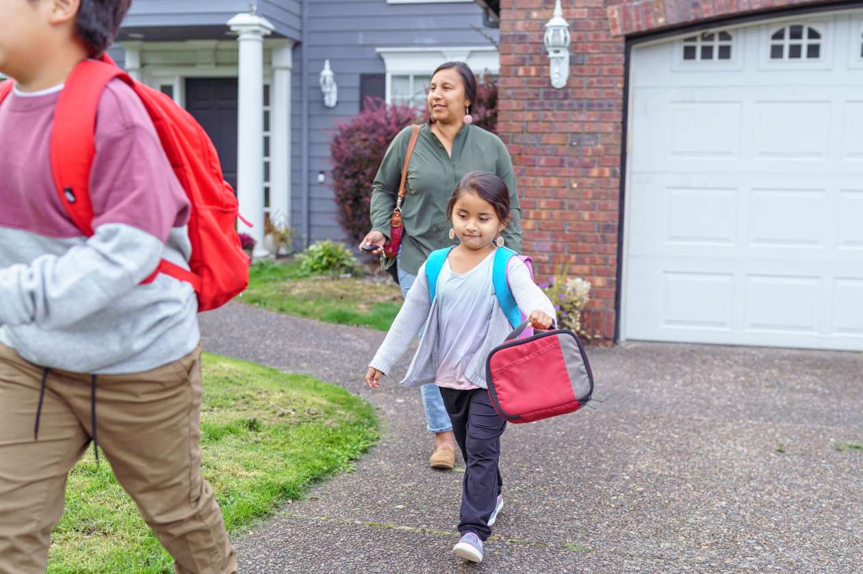 Mother and her son and daughter walk down the driveway. The chilldren wear backpacks and the daughter carries a lunchbox.