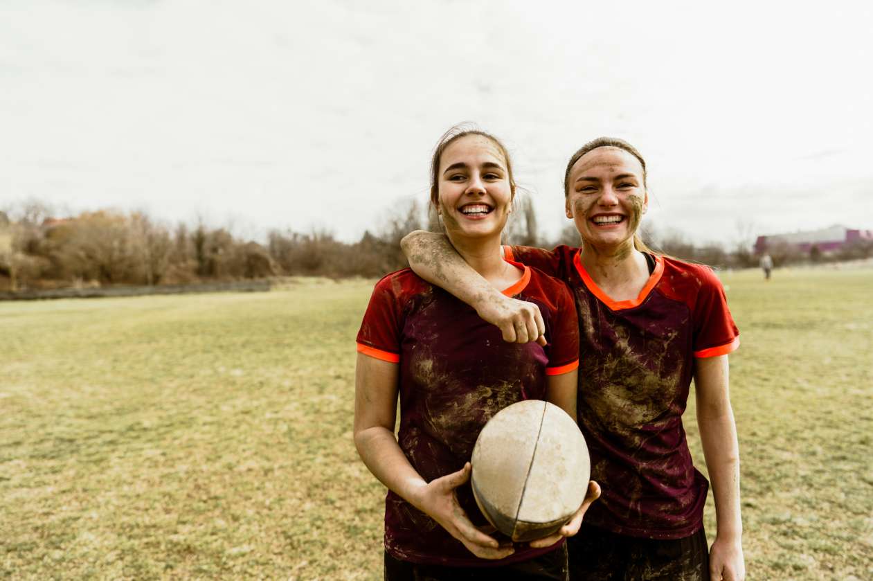 Two female rugby teammates stand on the field, smiling