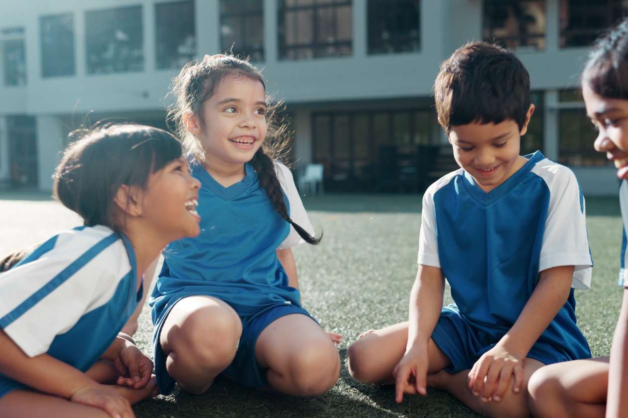 A group of kids wearing soccer jerseys sit on the field and laugh together.
