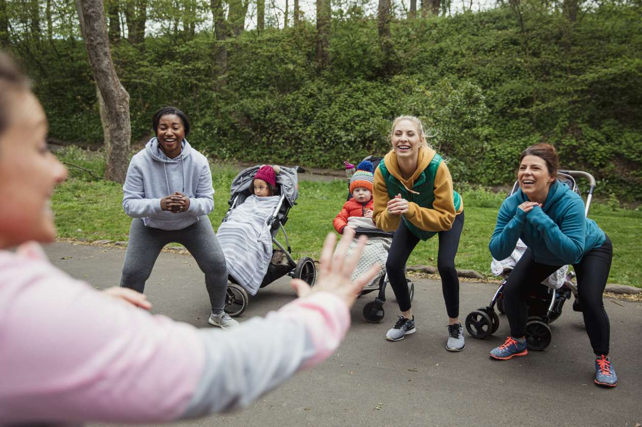 Moms do squats in an outdoor fitness class next to their babies in strollers.
