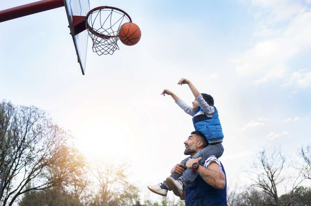 A son sits on his dad's shoulders and shoots a basketball on an outdoor court.