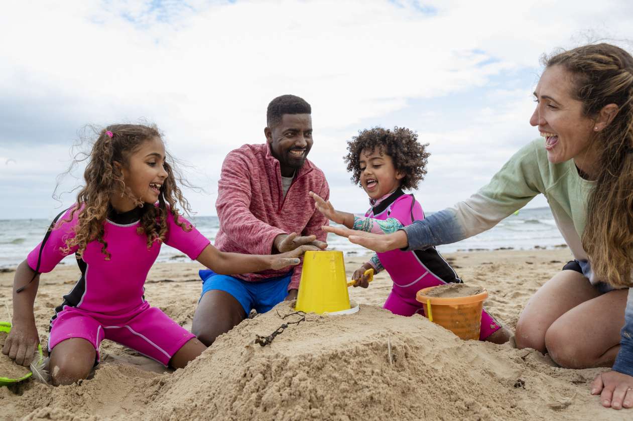 A father, mother, and their two daughters build a sandcastle on the beach.