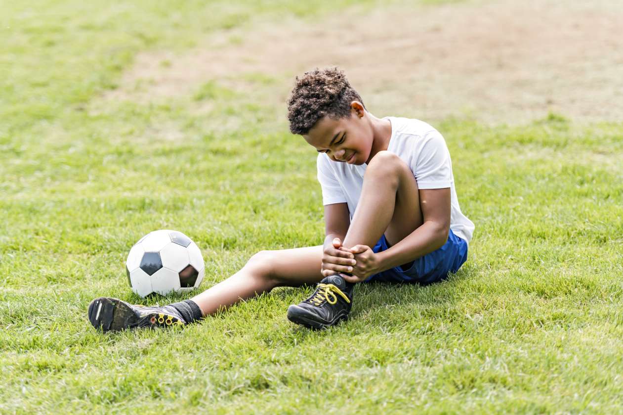 A boy sits on the soccer field, gripping his ankle and grimacing.
