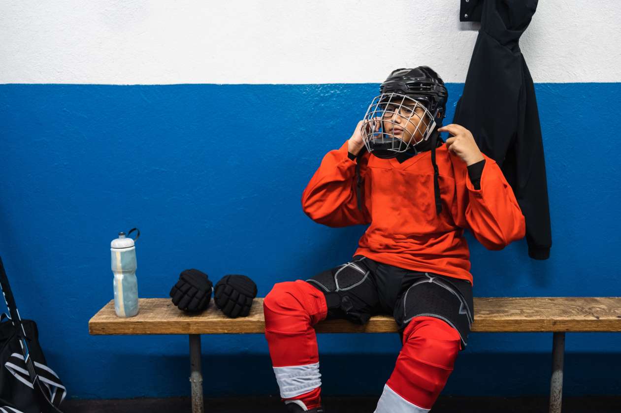 A young hockey player sits on the bench in the locker room and puts his helmet on.