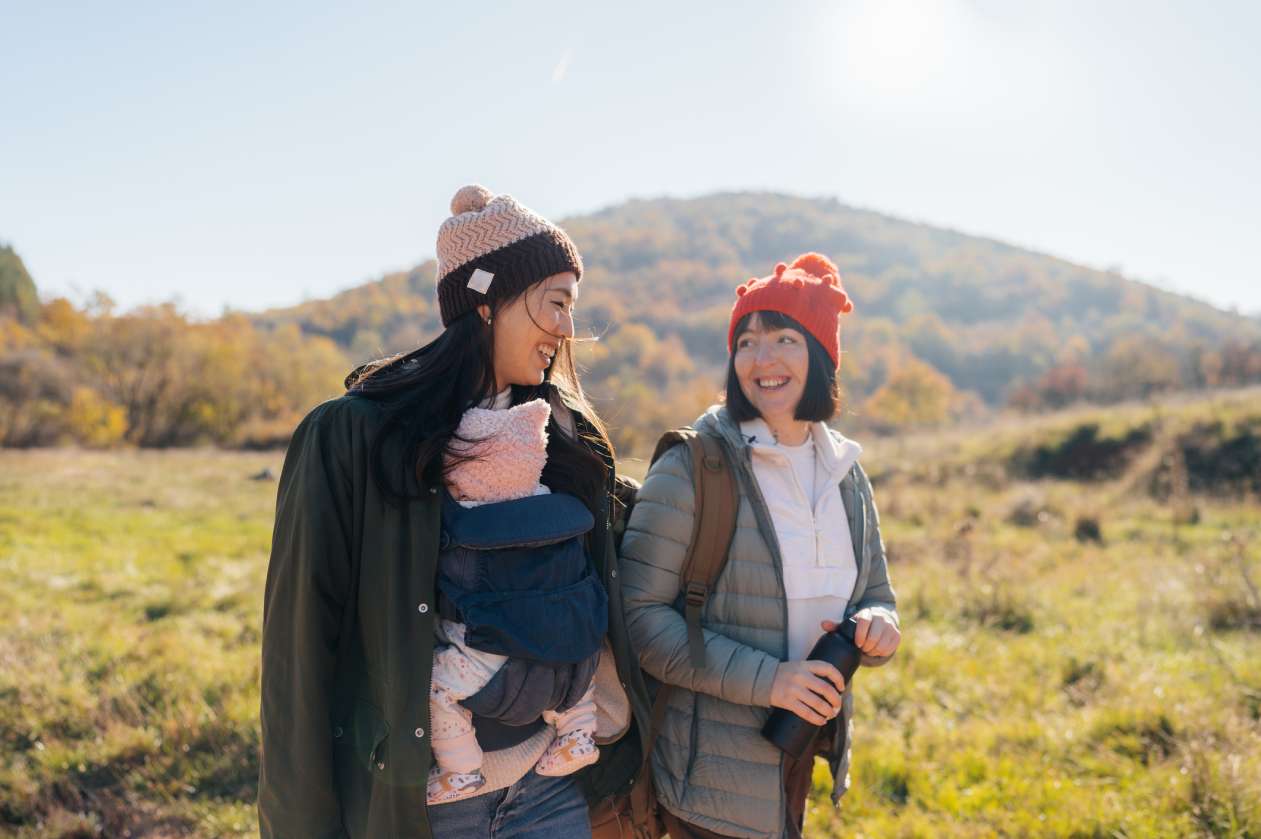 Two women walk together in the wilderness. One wears a baby in a carrier on her chest.