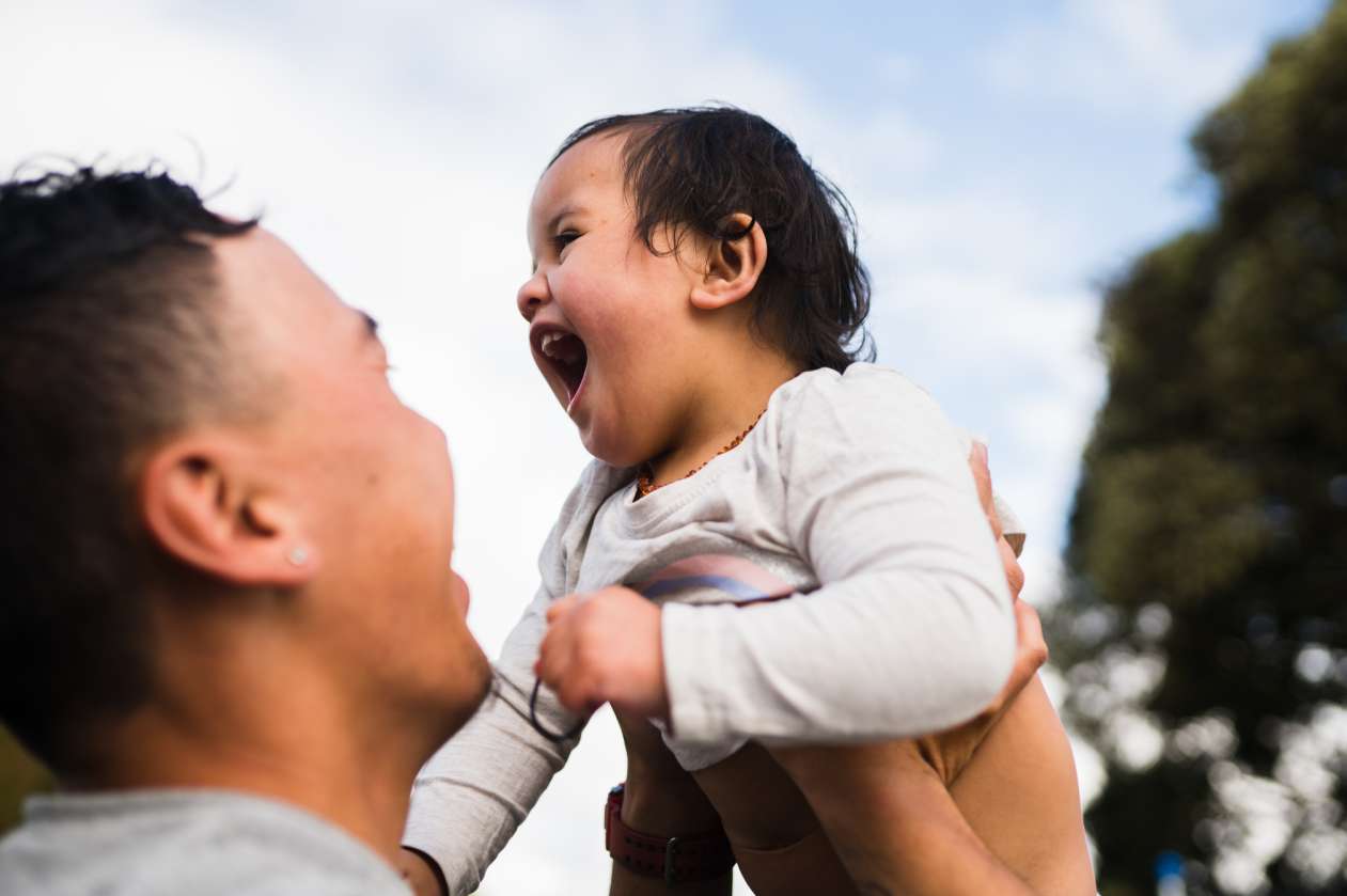 A dad holds his baby up in the air and smiles at him as the baby grins widely.