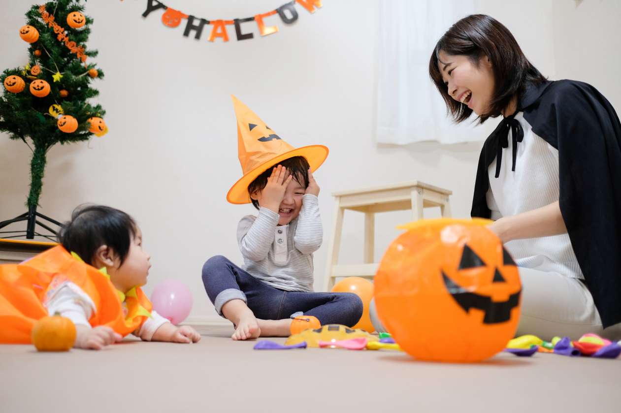A mom, her toddler, and her baby prepare for a Halloween party together. The toddler wears an orange witch hat while the baby wears a pumpkin costume.