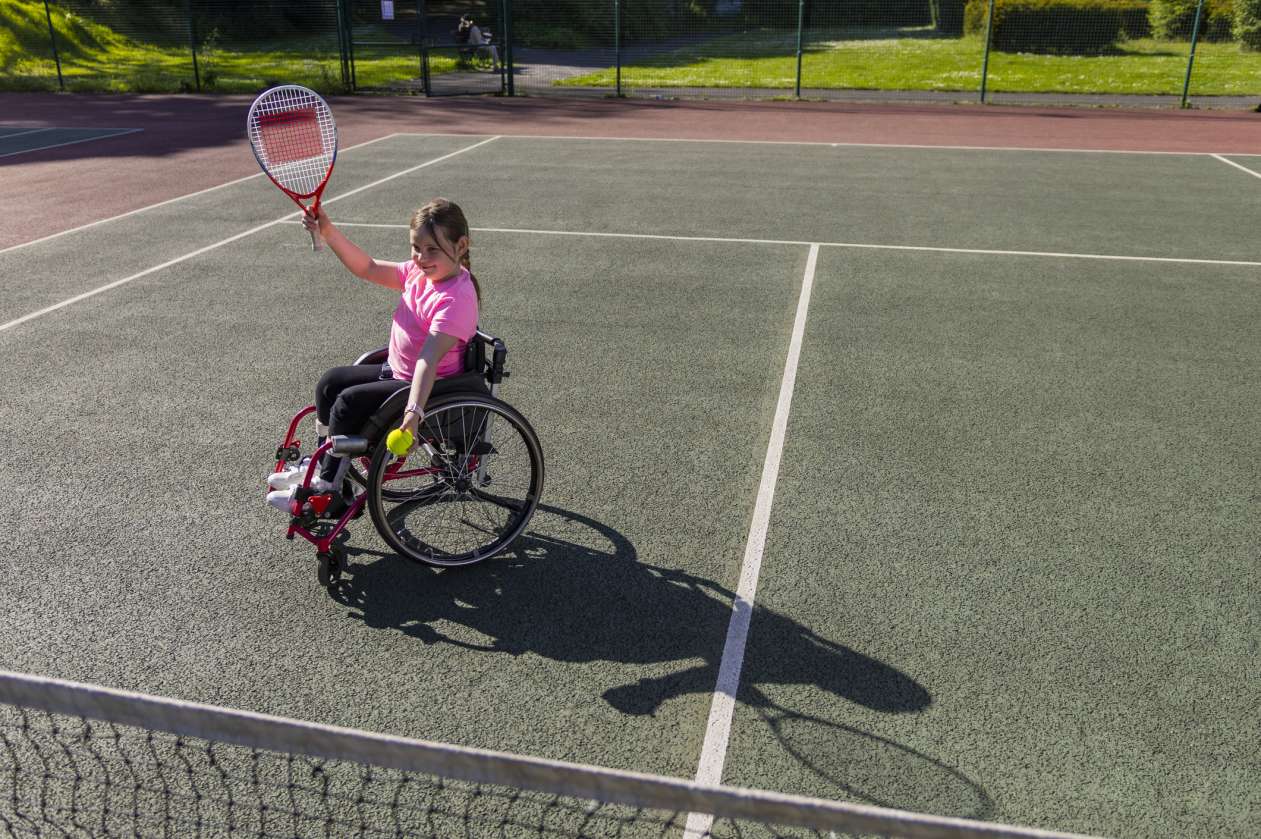 A young girl using a wheelchair prepares to serve a ball on an outdoor tennis court.