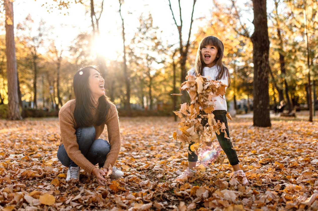 A mom and her young daughter toss leaves around outside on a sunny fall day.