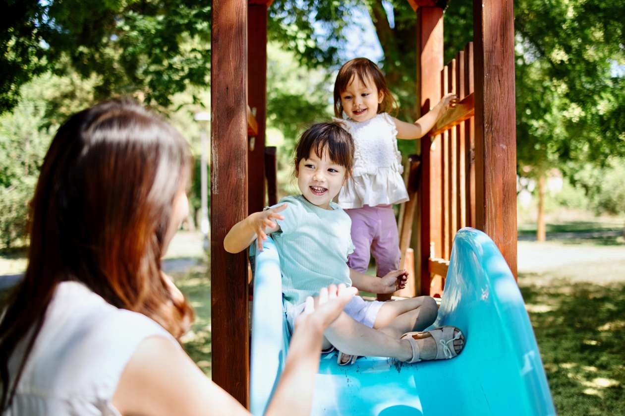 A childcare provider stands next to a playground slide as two young girls prepare to slide down it.