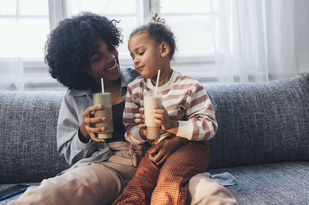 A mother and her young daughter sit on the couch in their living room together and drink smoothies.