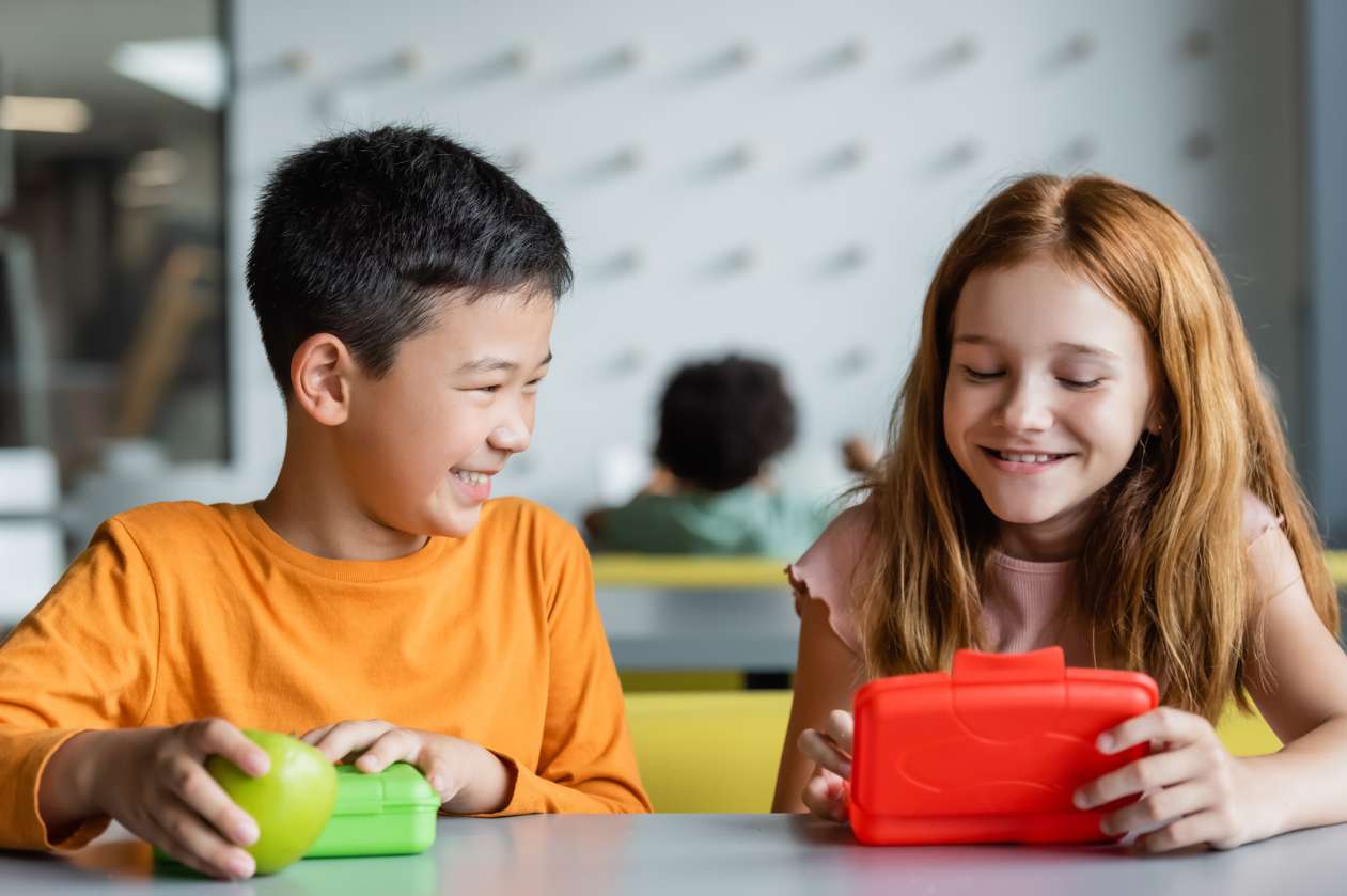 Two elementary students sit at their desk and take snacks out of their lunch boxes.