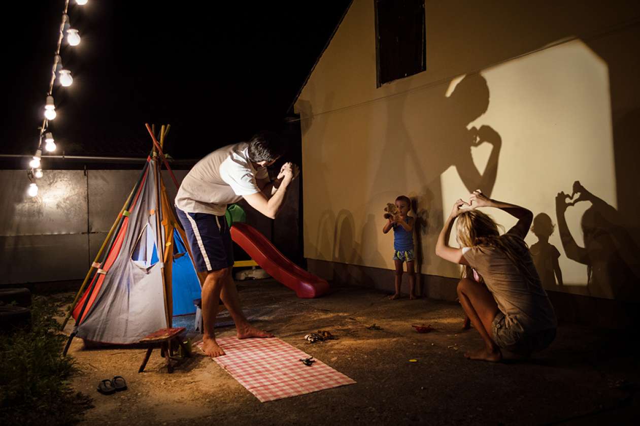 A father, mother, and their toddler make animal shadows with their hands  against a wall of their house.