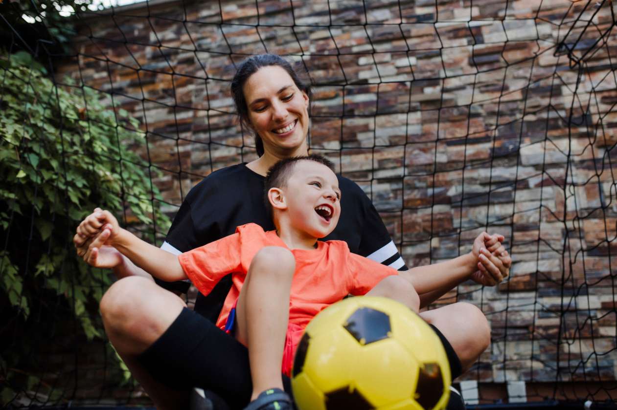 A mom and her son sit outside on the grass in front of a soccer net. The boy has a soccer ball in his lap.