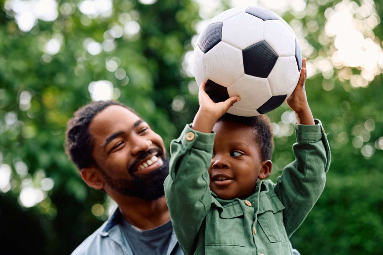 A father holds his young son in his arms and smiles at him. The son holds a soccer ball above his head.