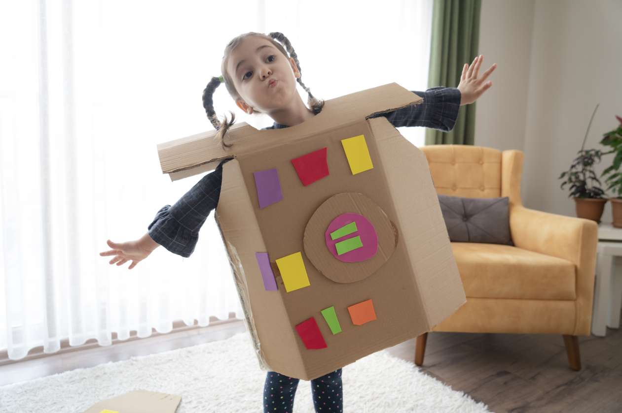 A girl stands in her living room wearing a cardboard box costume she has decorated with cut-outs of colourful construction paper.