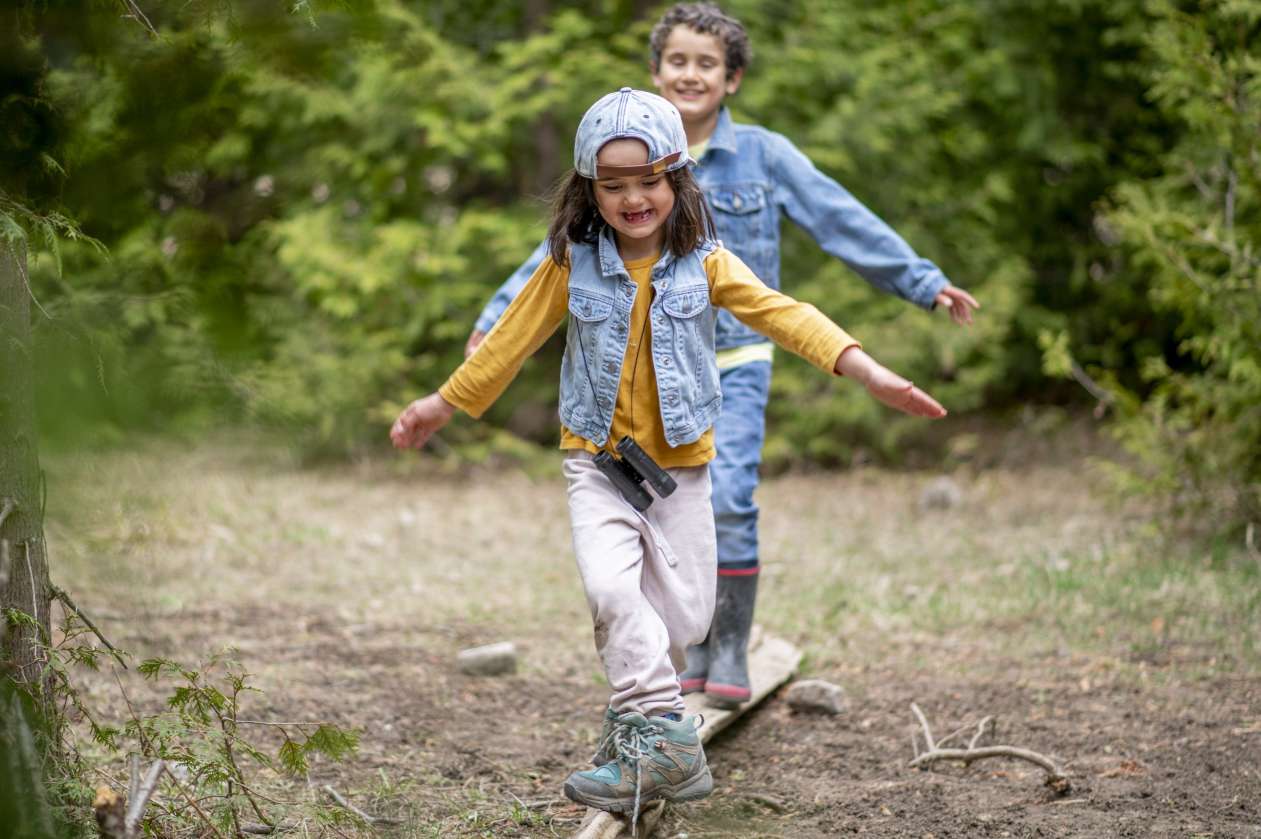 Two children balance on a log as they walk in the forest.