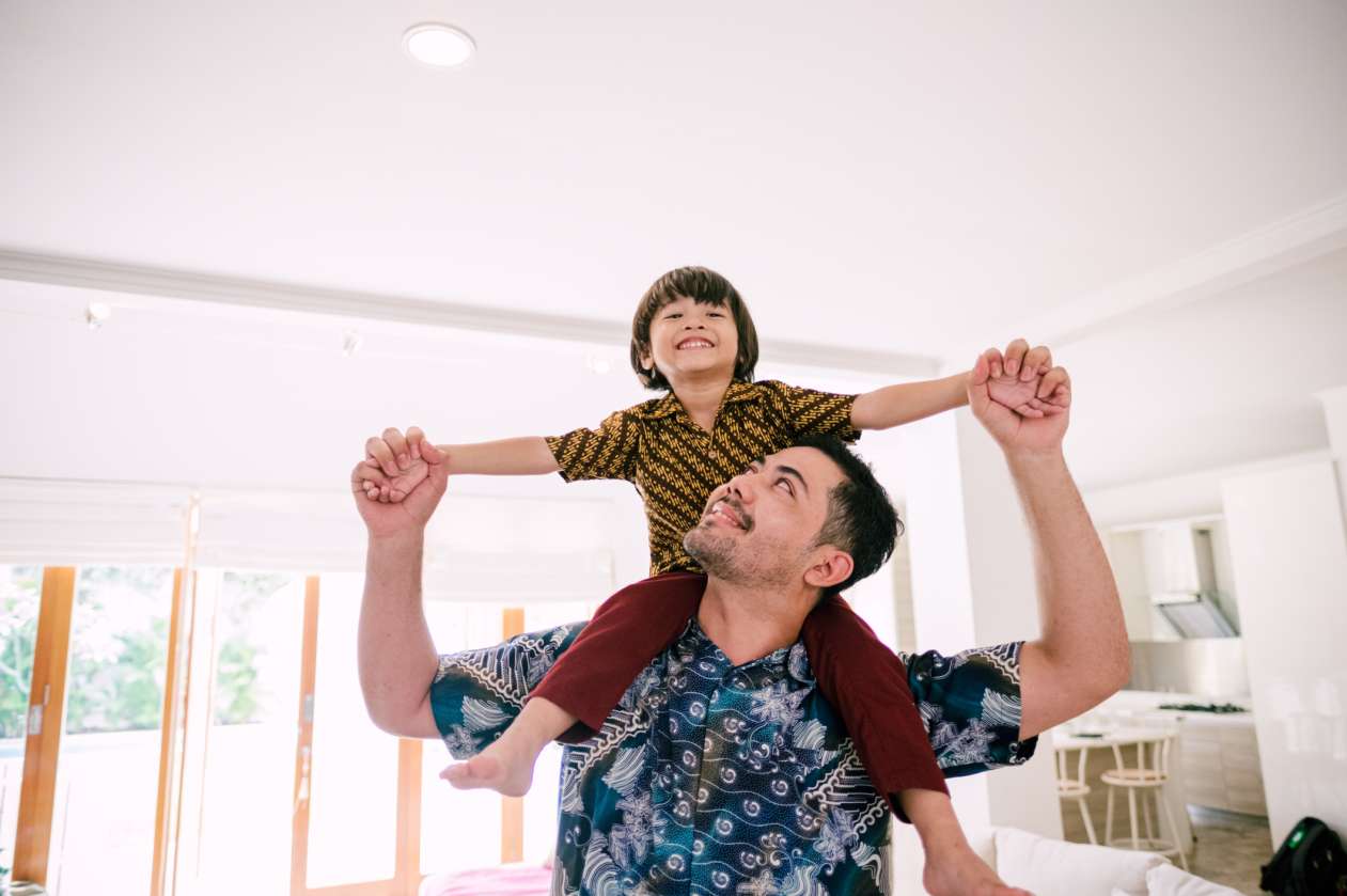 A young boy sits on his dad's shoulders with a smile on his face. They're in their living room at home.