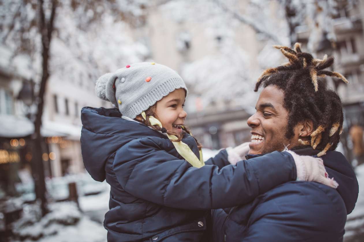 A father holds his daughter outside in a snowy park. Both are bundled up and they smile at each other.