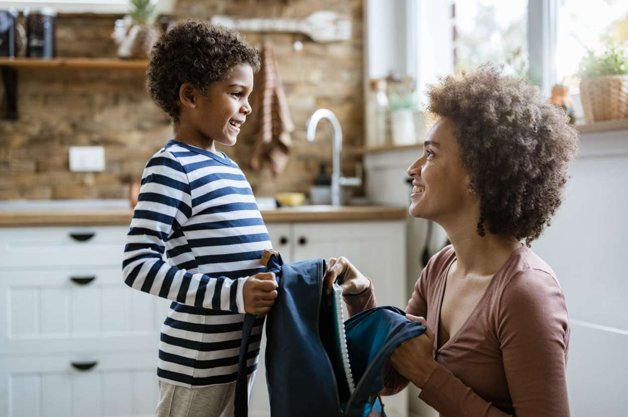 A mother and her young son stand in the kitchen, as the mom crouches down and hands her son his backpack. They're smiling at each other.