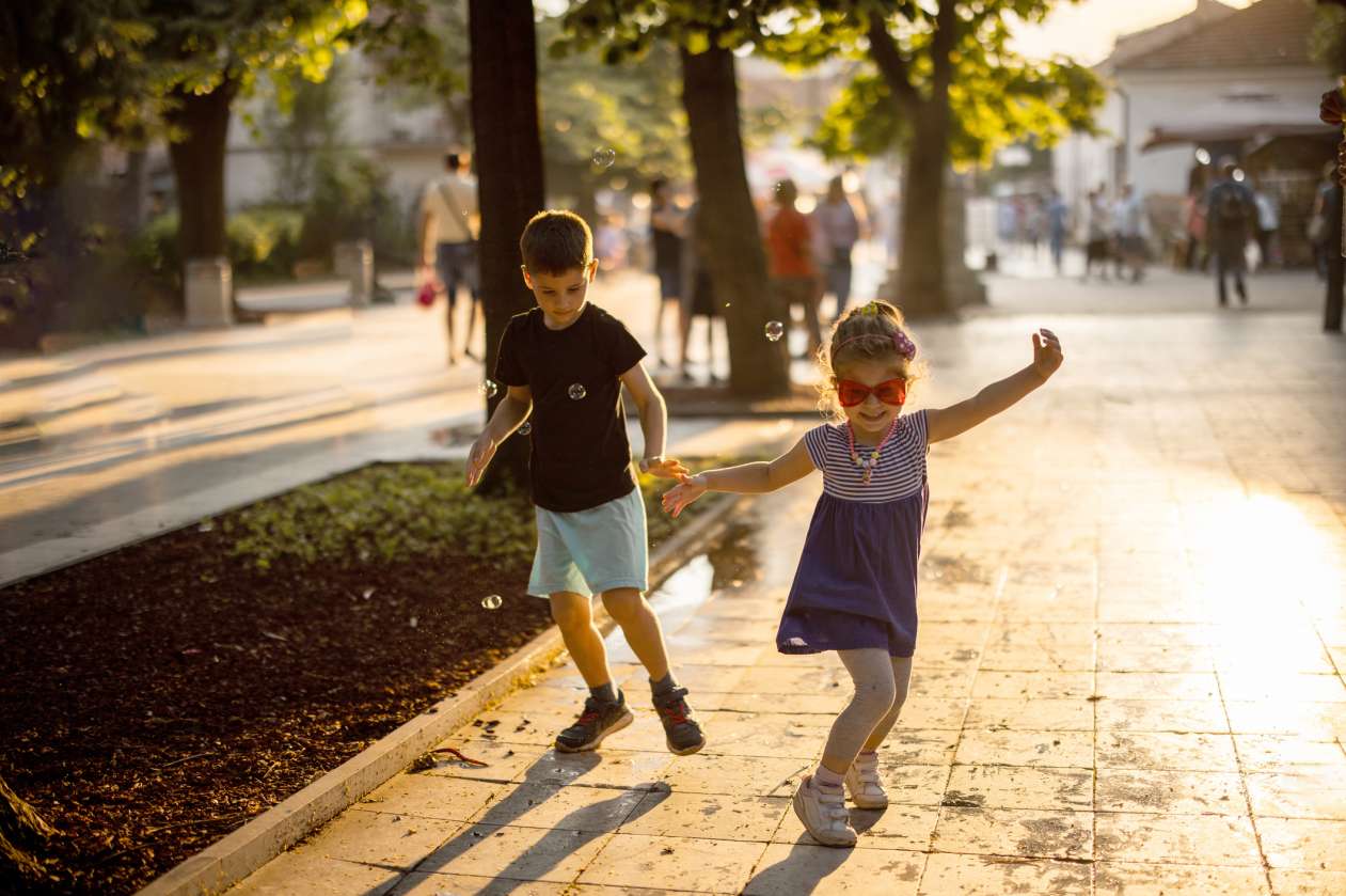 A brother and sister play and laugh on a city sidewalk.
