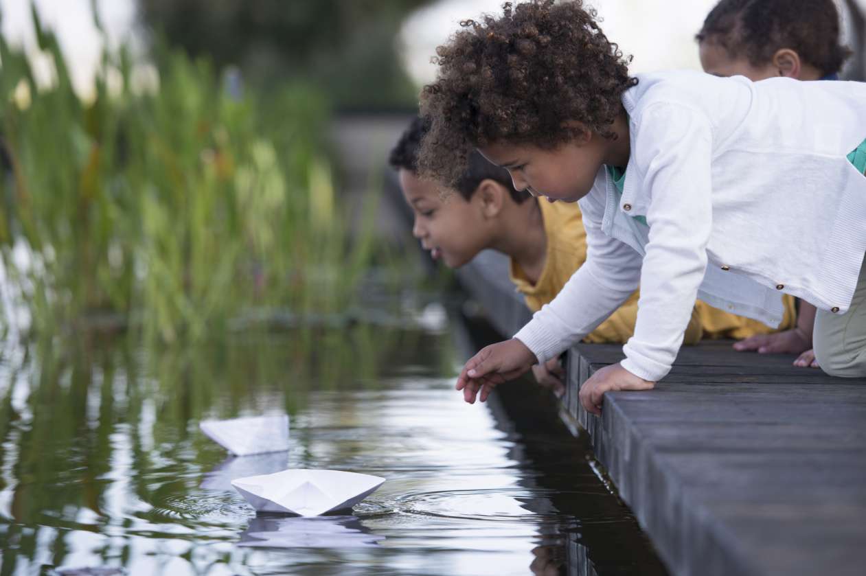 Two children crouch over a pond, having a race between two boats they made out of paper.