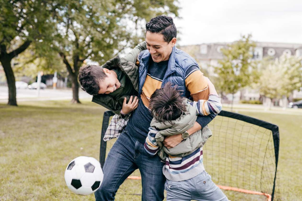 Dad plays soccer in the park with his two sons, all of them laughing